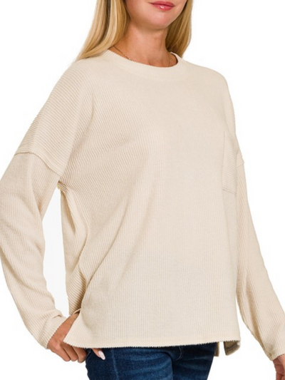 A woman stands confidently with one hand on her hip, wearing a loose-fitting sand beige Lindsey Ribbed Pocket Long Sleeve, effortlessly paired with blue jeans.