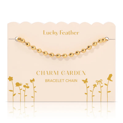 The Charm Garden Bracelet Chain - Gold is beautifully showcased on a cream-colored card featuring the words "Lucky Feather" and "Charm Garden Bracelet Chain." Its whimsical floral design and 14K gold plating make it a perfect personalized gift for those who admire artistic elegance.
