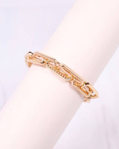 The Ursula Stretch Link Bracelet - Gold features textured rectangular links, perfect for stacking and displayed on a white cylindrical stand.