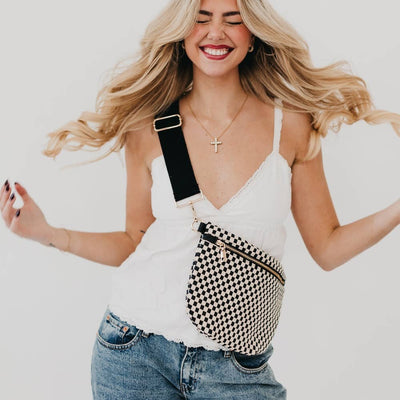 A smiling woman with long, wavy blonde hair, eyes closed, wears a white tank top and blue jeans while posing with the Westlyn Woven Bum Bag in Checkered Black across her chest.