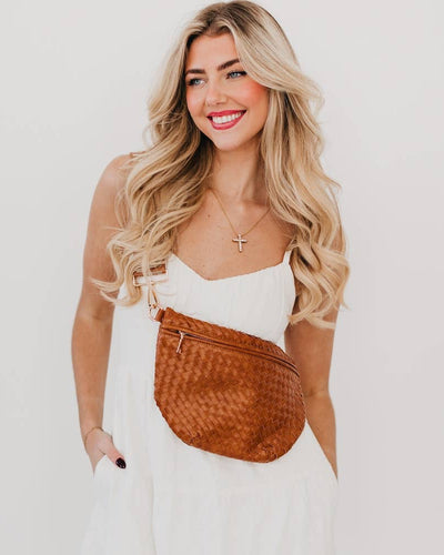 A smiling woman with long blond hair wears a white dress and the Westlyn Woven Bum Bag - Brown in vegan leather with an adjustable strap, standing against a plain light background with one hand in her pocket.
