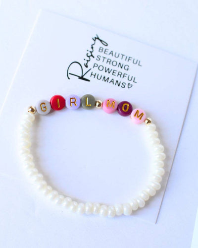 The Girl Mom Beaded Stretch Pearl Bracelet in multi-color features pearl beads and colorful letters spelling "GIRL MOM." It comes on a card that says, "Raising Beautiful Strong Powerful Humans," capturing its essence.