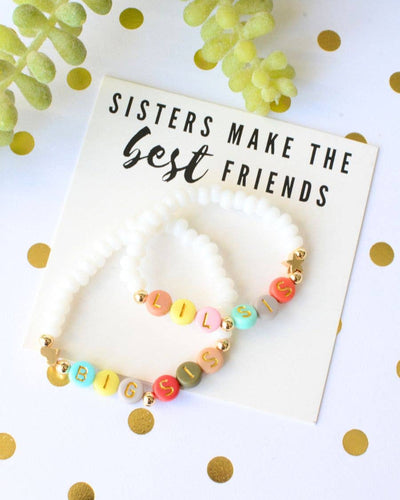 The "Big Sis Lil Sis Best Friends Bracelets - Pearl / Multi Colored" set includes two beaded bracelets with letter beads spelling "LIL SIS" and "BIG SIS," displayed on a card reading, "Sisters make the best friends," with gold polka dots and green plant accents.