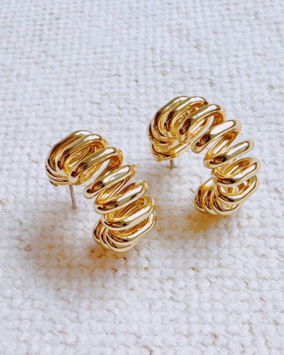 The Curly Cord Hoop Earrings feature a chunky gold, layered spiral design and are shown on a textured off-white fabric background.