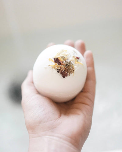 A hand holds a Bath Bomb - Lavender enriched with shea butter, topped with dried herbs and flowers. The blurred background suggests a calming bathroom setting, ideal for aromatherapy.