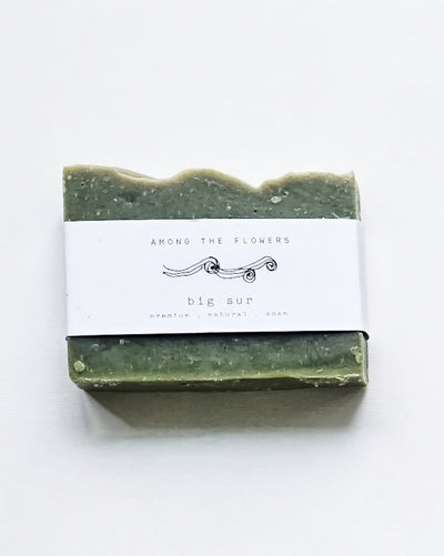 A handcrafted Big Sur soap bar wrapped in a white label on a white background, featuring "Among the Flowers" and "Big Sur" text, highlighting its premium natural ingredients and decorative lines. Product: Cold Processed Soap - Big Sur.