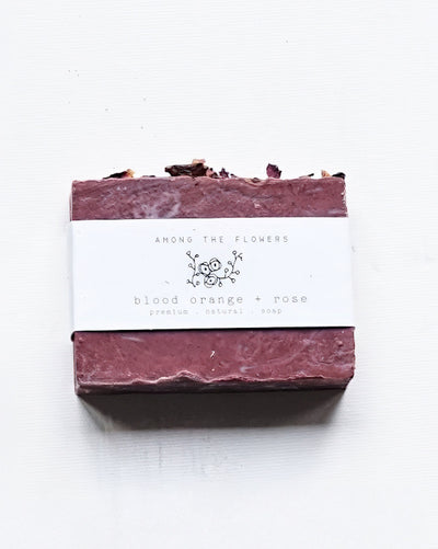 A deep red rectangular bar of "Cold Processed Soap - Blood Orange + Rose" wrapped in a white label reading "AMONG THE FLOWERS" and "premium, natural soap." The handmade soap features dried flowers embedded on top.