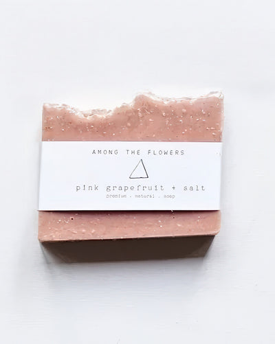 The Cold Processed Soap - Pink Grapefruit + Salt features a jagged top and is wrapped in a white label reading "AMONG THE FLOWERS" with notes of "pink grapefruit + salt." Infused with Himalayan Pink Salt, it rests on a white backdrop highlighting its organic beauty.