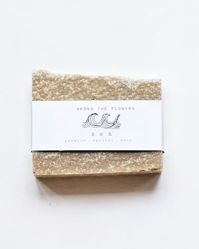 A rectangular beige bar labeled with waves reads "Among the Flowers, Cold Processed Soap - Sea, fragile natural rouge," is textured like stone and infused with mineral-rich sea salt.