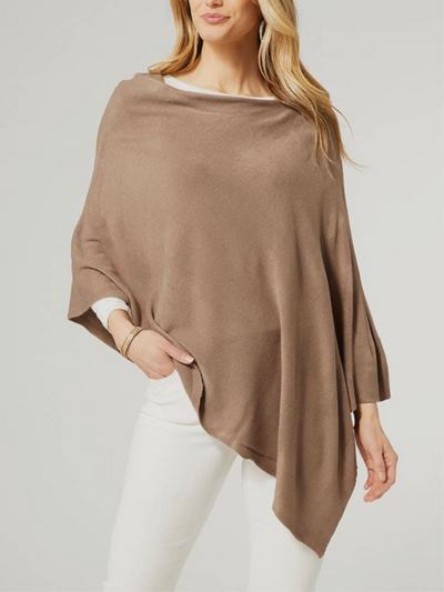 A person showcases the Kayley Lightweight Poncho in Mushroom, elegantly layered over a white top and pants. They stand stylishly with one hand in their pocket against a plain background, highlighting the poncho's versatile appeal for fashion and as a gift.