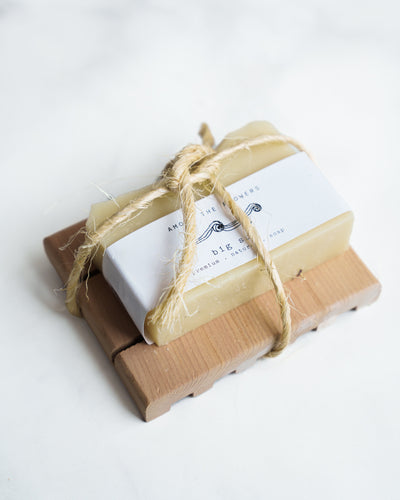 The Soap + Deck Bundle - Big Sur includes an exfoliating soap wrapped in white paper with a black design, tied with rustic rope, and placed on a wooden dish, showcasing its natural ingredients against a light backdrop.