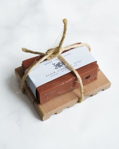 The Soap + Deck Bundle - Blood Orange + Rose features a handmade blood orange soap with rose clay, wrapped in white paper and twine, on a Redwood Soap Deck. The reddish-brown bar is labeled "blood orange rose," with floral graphics on a pristine white backdrop.