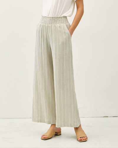 A person wears Calla Pinstriped Gauze Pants in sage—a light, wide-leg, relaxed-fit style—paired with a white sleeveless top and tan open-toe sandals, standing against a plain white background with hands in pockets.