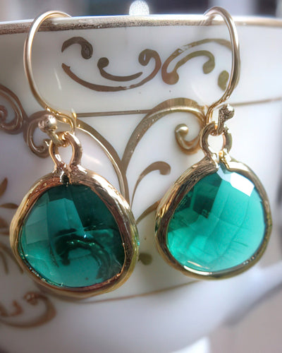 A pair of elegant teardrop earrings featuring 15x18.5mm faceted green glass gems set in gold-plated bezels with 14k gold-filled earwires. The earrings are displayed hanging on a decorative white ceramic dish with gold swirls, showcasing their refined and luxurious design.