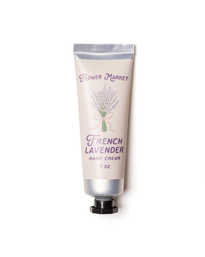 A product image of the Flower Market French Lavender Hand Cream in a 1 oz tube. The packaging features a soft cream-colored background with a lavender bouquet illustration tied with a pastel ribbon. Text reads "French Lavender Hand Cream" in purple, along with "Flower Market" at the top. The tube has a silver cap and an elegant design, ideal for gifting or daily use.