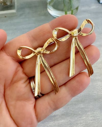 A pair of gold stud bow earrings measuring approximately 1.5 inches, with a shiny and textured finish, displayed resting on a hand. These earrings are perfect for gifting or adding a touch of elegance to everyday outfits.