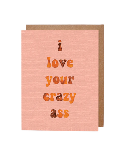 A peach-toned greeting card with subtle textured detailing featuring the phrase "i love your crazy ass" written in a playful, orange and brown gradient font. The card is accompanied by a brown kraft envelope in the background.