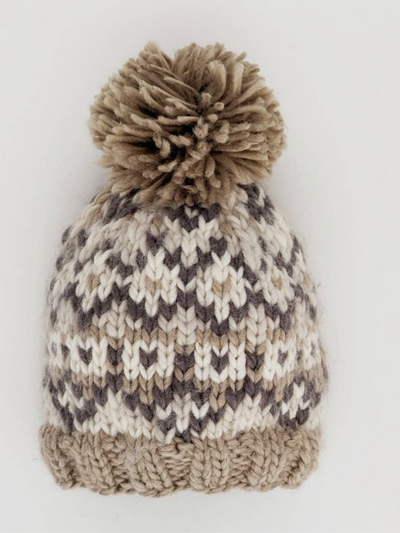 Introducing the Littles Fairisle Beanie - Pebble, a cozy winter hat made from chunky yarn in beige, white, and gray. It features a large, fluffy pompom and is crafted from recycled polyester sourced from plastic bottles. The beanie is beautifully set against a plain white background.