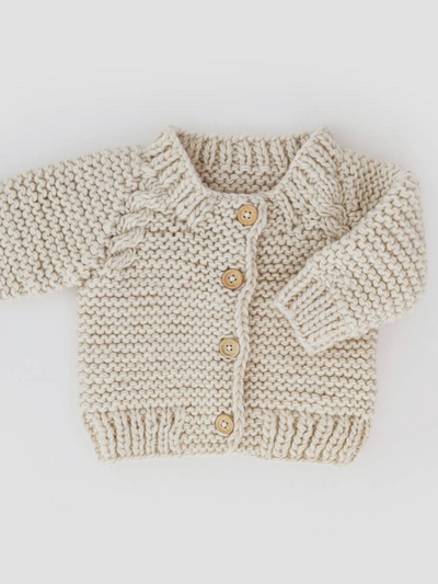 The Littles Garter Stitch Cardigan Sweater in Natural, featuring wooden buttons and a textured garter stitch with ribbed cuffs, hem, and collar, elegantly rests on a white background. It's an adorable hand-knitted addition to any baby and toddler clothing collection.