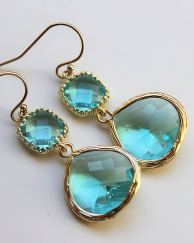 A pair of stunning aquamarine drop earrings featuring 15x18.5mm teardrop gems and 9mm square gems, set in gold-plated bezels with 14k gold-filled earwires. Displayed on a white background, these earrings exude elegance and vibrant charm.