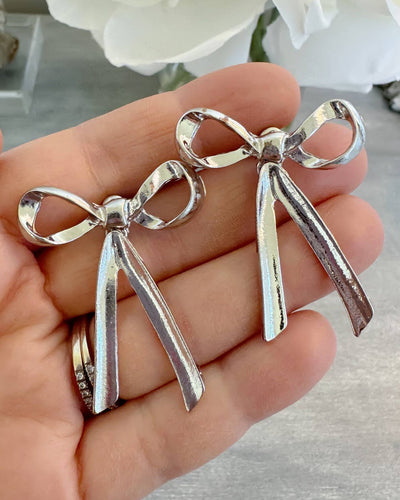 A pair of silver stud bow earrings measuring approximately 1.5 inches, with a shiny and polished finish, displayed resting on a hand. These earrings are perfect for gifting or as a stylish addition to everyday outfits.