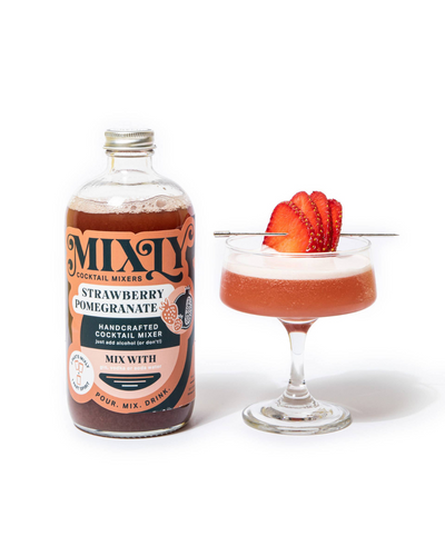 A bottle of Mixly Strawberry Pomegranate Mixer sits beside a coupe glass with a pink cocktail, garnished with sliced strawberries—this handcrafted mixer is perfect for creating both cocktails and mocktails.