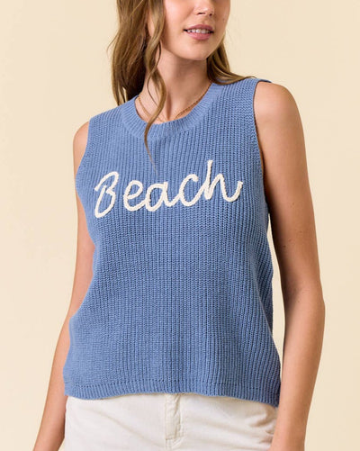 Woman wearing the "Beach" Round Neck Tank Top in light blue, featuring white BEACH script embroidery and a ribbed knit texture, paired with light shorts and standing against a beige background.