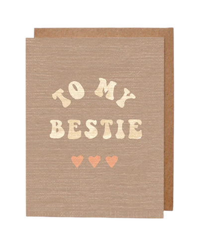 A beige greeting card with a subtle textured design featuring the phrase "TO MY BESTIE" written in gold letters with a playful font. Below the text are three small pink hearts in a row. A brown kraft envelope is partially visible in the background.