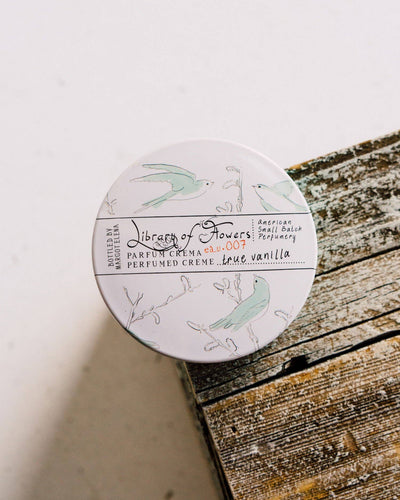Library of Flowers True Vanilla Parfum Crema tin resting on a rustic wooden surface. The lid displays an intricate botanical illustration with light blue birds and branches, emphasizing the perfume's elegant and artistic packaging.