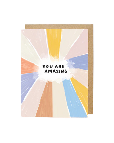 A greeting card with a colorful abstract design featuring pastel rays in shades of orange, yellow, pink, blue, and cream radiating outward. In the center, a white cloud shape contains the bold, black text "YOU ARE AMAZING." The card is paired with a brown kraft envelope in the background.