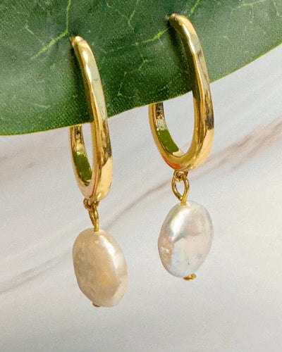 Dangle Pearl Gold Hoop Earrings feature dangling oval freshwater pearls, shown hanging from a green leaf on a marble background—a striking jewelry collection addition.
