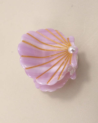 A lilac Ocean Cellulose Seashell Hair Claw Clip with gold lines and a pearl accent rests on a beige surface, resembling a delicate hair accessory.