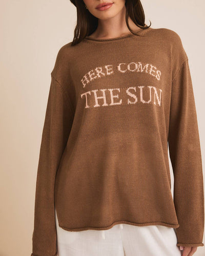 A plus-size model with dark hair wears the "Here Comes The Sun" Graphic Sweater in Cocoa. Against a neutral background, they pair it with white pants, showcasing effortless style.