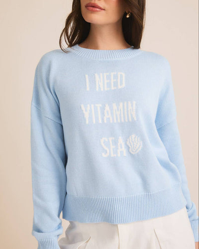Wearing an "I NEED VITAMIN SEA" Sweater in baby blue with a seashell graphic, a woman sets off her long dark hair with a white skirt against a neutral backdrop, ideal for beach lovers.