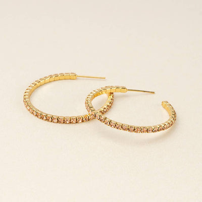 The Sparkle & Shine Sm Rhinestone Hoop Earrings in Champagne and Gold glisten with embedded sparkly stones as they elegantly rest on a light beige surface. Crafted from 14K gold-dipped brass, these open-ended, textured earrings offer a luxurious touch while being hypoallergenic for sensitive ears.