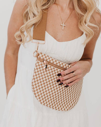 A woman with wavy blonde hair wears a gold cross necklace and holds the Westlyn Woven Bum Bag in Checkered Tan, crafted from vegan leather. Her nails are dark and manicured, with one hand casually slipped into her pocket.