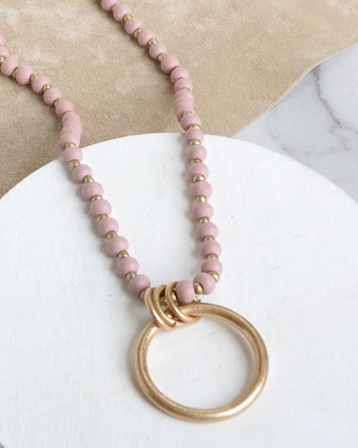 The Long Wooden Bead Necklace with Gold Circle - Pink features boho-chic pink wooden beads, tiny gold spacers, and a standout gold ring pendant with three smaller rings, elegantly displayed on a white and beige background.
