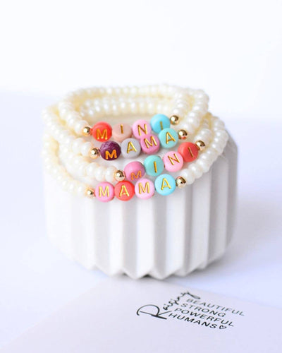 The "Mama and Mini" bead bracelets, from the Pearl/Multi Colored collection, feature white and gold beads with colorful "MINI" and "MAMA" letters. These elastic designs elegantly rest on a white fluted ceramic holder, with a partial text note visible nearby.