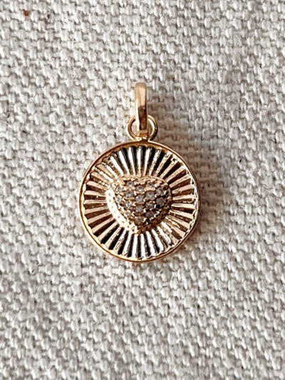 The 18K Gold Filled Spoke Textured Charm Heart features a heart design with sunray-like patterns, crafted from hypoallergenic 18k gold filled material, set on a textured fabric background.
