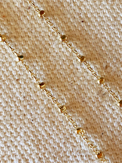 A close-up of the 18K Gold Filled 1mm Spaced Beaded Chain on textured beige fabric highlights its delicate and elegant design. The chic chain, with evenly spaced round beads, offers hypoallergenic comfort that contrasts beautifully with the fabric's weave.