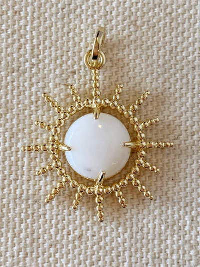 The 18K Gold Filled Radial Sun Pendant is handcrafted in Brazil, featuring a sun-shaped gold design with a central white stone on a textured beige background and intricate sunray details radiating from the stone.