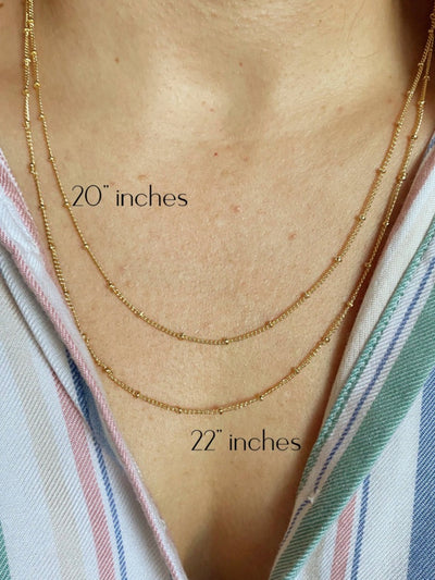 A person wears two delicate 18K gold-filled, hypoallergenic satellite chain necklaces over a striped shirt; the top one is "20 inches" and the bottom is "22 inches," offering a stylish yet comfortable look.