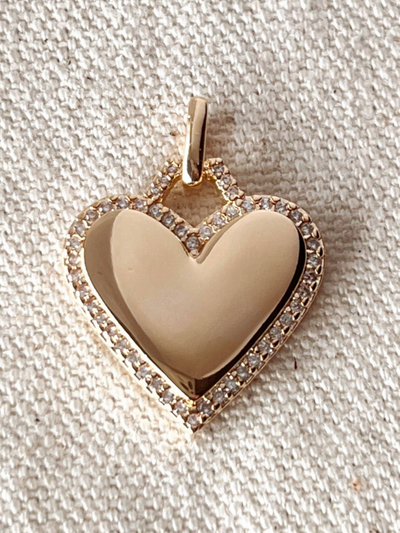 An 18K Gold Filled Heart Pendant with a smooth center and cubic zirconia-studded edges rests on a textured, off-white fabric background.