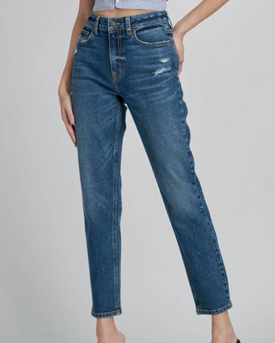 A person wears vintage-style High Rise Slim Straight dark denim, featuring faded patches and slight distressing on the upper thighs. They're paired with black heels, and the person poses with one hand on their hip.