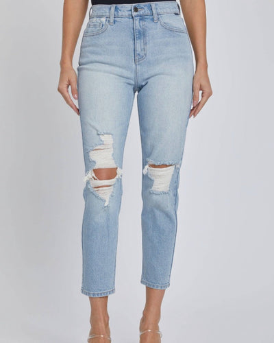 A person stands against a plain light background wearing the High Rise Mom Crop Jean - Light Denim featuring distressed details and large knee rips, styled with clear heels.