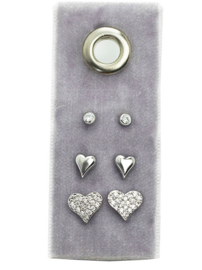 The Heart Velvet Stud Gift Set - Silver includes three pairs of earrings on a gray card: small round studs, smooth silver heart-shaped studs, and larger hearts with sparkling cubic zirconia. Crafted in elegant plated brass for a glamorous touch.
