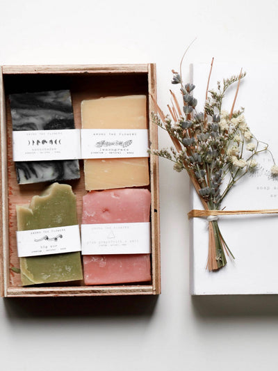The Soap Sampler Set features a wooden box containing four premium soap bars, each infused with essential oils and in elegant shades: black and white, beige, green, and pink. Beside it is a twine-bound white book adorned with dried flowers.
