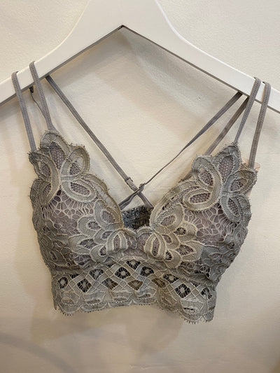 The Ava Lace Bralette in Sleet, featuring intricate floral crochet lace patterns, hangs on a white clothes hanger against a light background. It boasts adjustable straps that elegantly cross at the back for enhanced comfort and style.