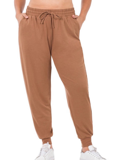 A person lounging comfortably in the Carmen Jogger Pants - Cocoa, with hands in pockets, paired stylishly with white sneakers against a plain white background.